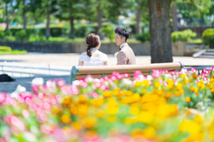 黄やピンクのカラフルな花が咲く花壇の前で笑いながら会話をする新郎新婦