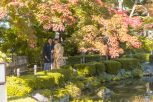 赤く色づく庭園で金色の色打掛に身を包む新婦と紋付袴姿の新郎